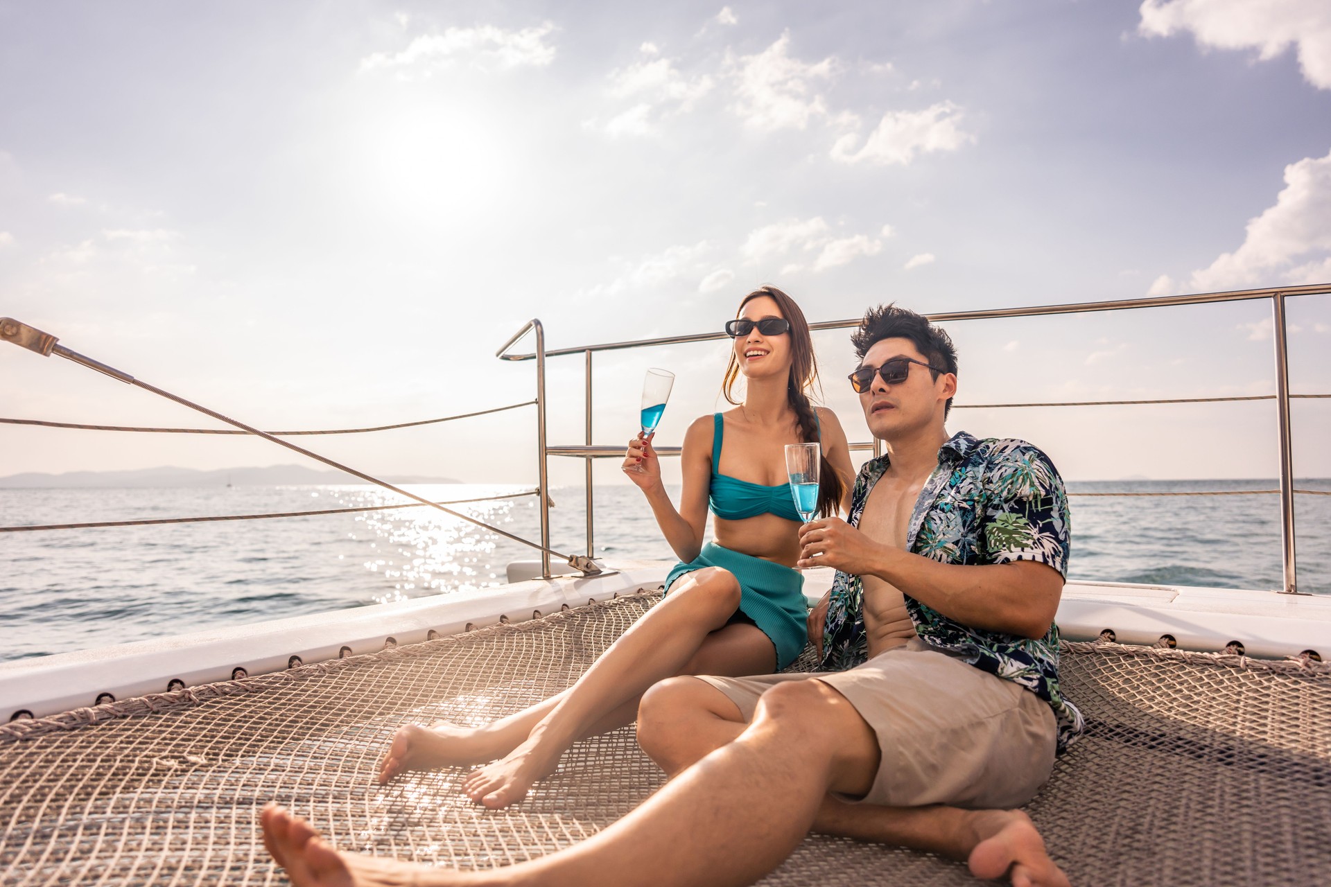 Asian beautiful couple drinking champagne while having party in yacht. Attractive man and woman hanging out, celebrating anniversary honeymoon trip while catamaran boat sailing during summer sunset.